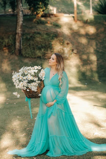 Pregnant woman in a teal dress holding a basket of flowers in a sunlit garden setting.