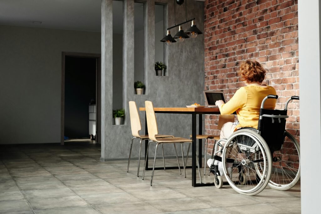 A woman in a wheelchair working on a laptop at home with a brick wall backdrop.