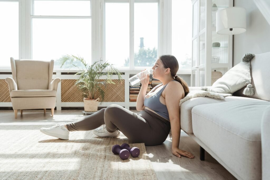 Curvy woman in activewear, drinking water in a sunny living room with dumbbells nearby.